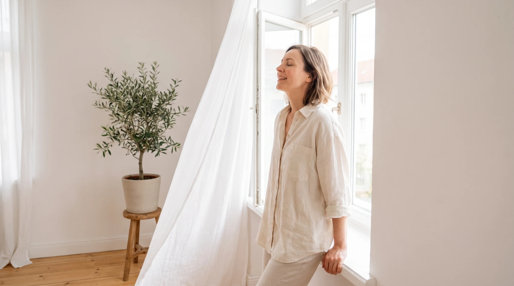 Mujer respirando tranquila junto a una ventana abierta con la cortina al viento en una habitación luminosa
